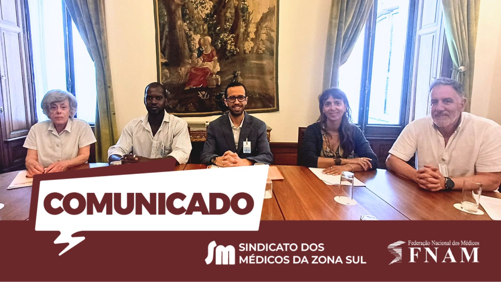 Fotografia de uma reunião institucional com cinco pessoas sentadas lado a lado atrás de uma mesa comprida de madeira. No centro da mesa está André Arraia Gomes, presidente da FNAM, ladeado por outros quatro representantes (duas mulheres e dois homens), todos vestidos com roupa casual-formal. Estão num salão clássico com janelas altas e uma grande tapeçaria antiga pendurada na parede ao fundo. A parte inferior da imagem é sobreposta por uma faixa horizontal de cor vinho, que exibe um balão de diálogo branco à esquerda com a palavra 